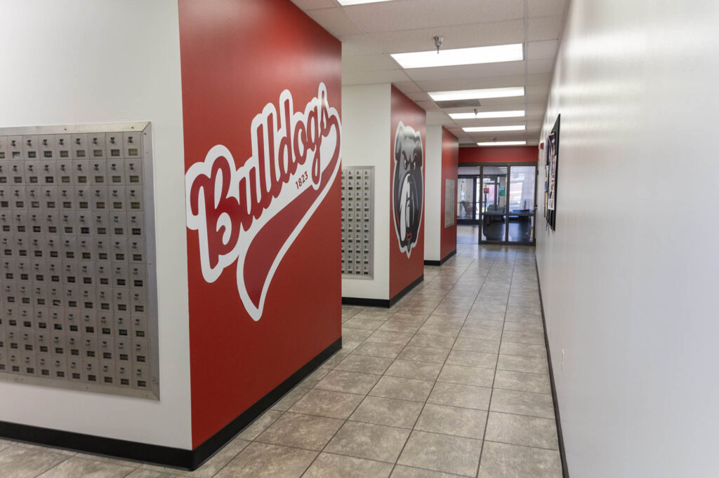 A hallway with red and white walls, Bulldogs painted on the left wall, mailboxes on the left, and a bulldog mural visible ahead. The floor is tiled, and there are overhead fluorescent lights.