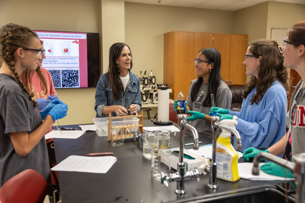 A teacher smiles while instructing four students wearing safety goggles and blue gloves at a laboratory table with beakers and a test tube rack.