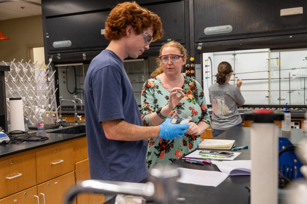 A student and instructor in safety goggles discuss a beaker in a laboratory while another person works in the background.