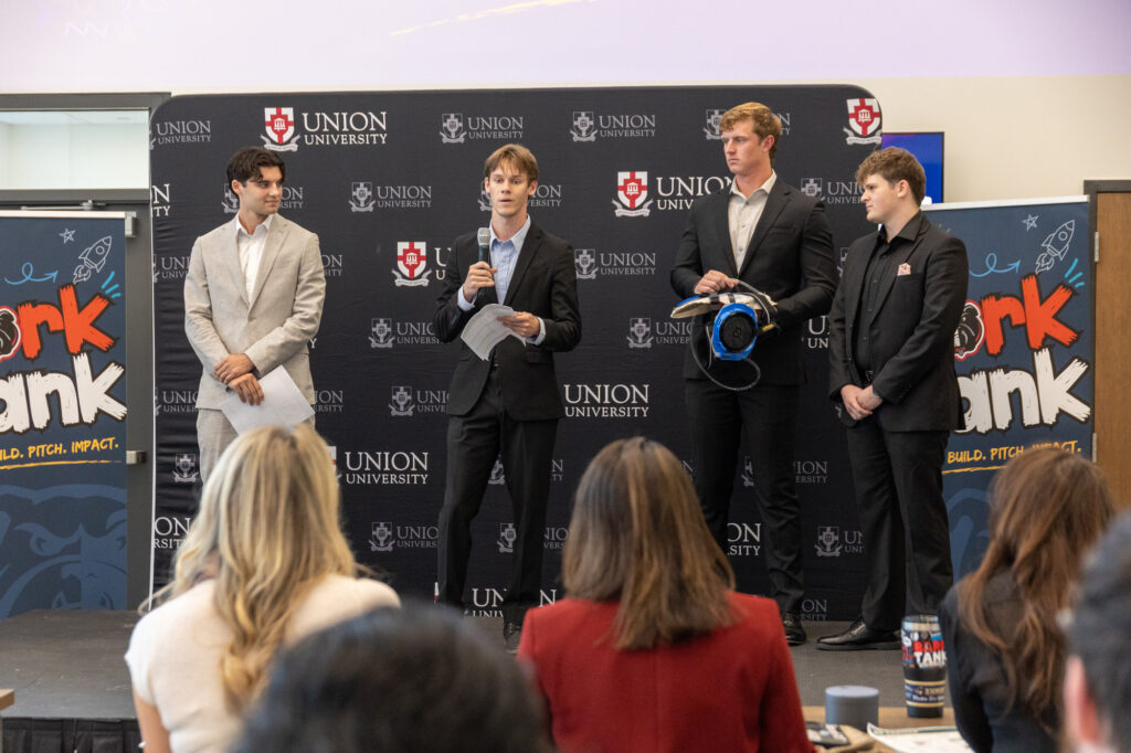 Four men stand on a stage in front of a Union University backdrop; one man speaks into a microphone while holding papers, and another holds a blue protective helmet.