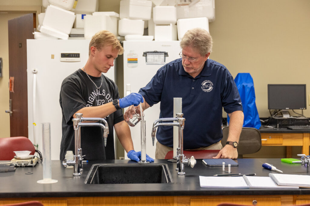 A student wearing blue gloves pours liquid from a beaker into a graduated cylinder held by an instructor in a laboratory.