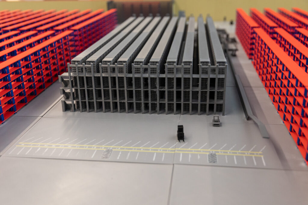 A scale model of a warehouse facility featuring a central gray storage structure flanked by tall red shelving units and a parking lot.