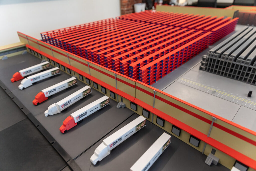 A scale model of a warehouse facility featuring rows of red storage racks and multiple white Tractor Supply Co. semi-trucks parked at loading docks.