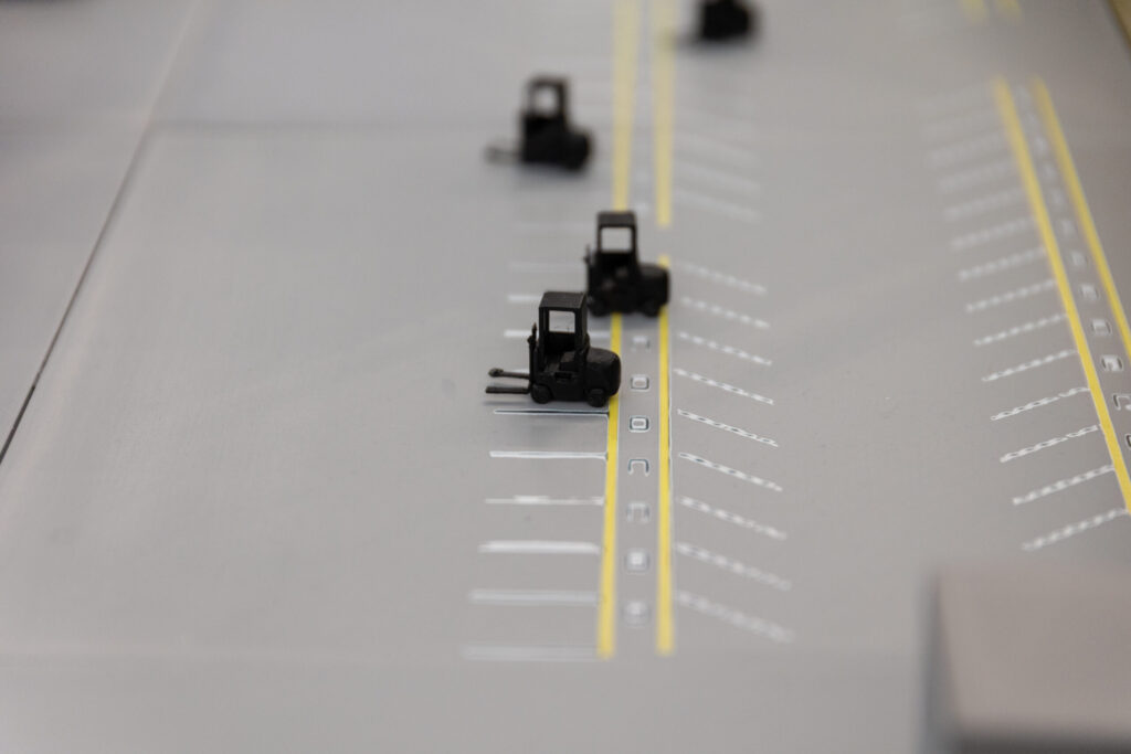 Three miniature toy forklifts positioned on a model warehouse floor with yellow aisle markings.