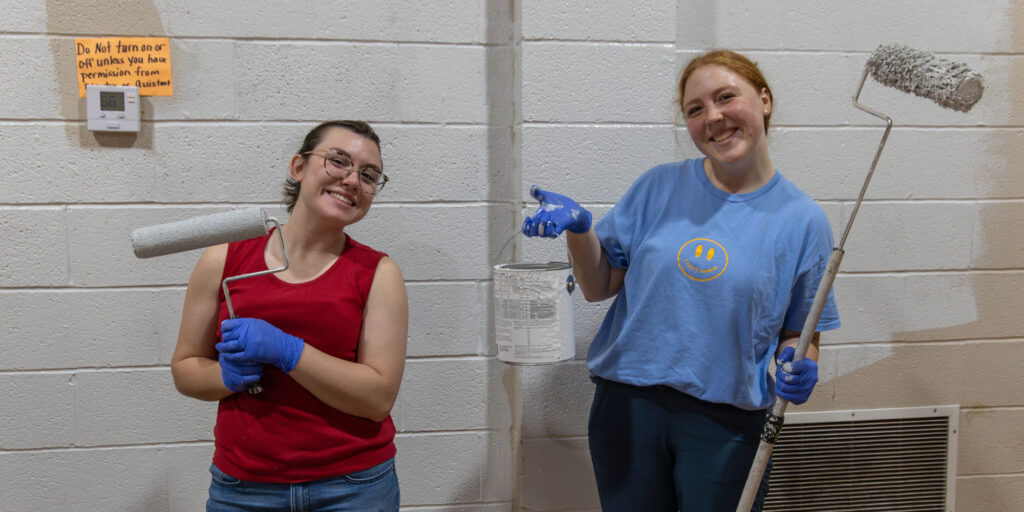 Two people holding paint rollers and a paint can stand in front of a white brick wall during a Union University Campus and Community Day.