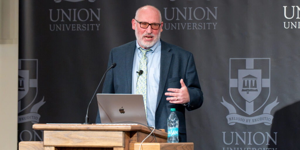 Steve Pincus speaks at a wooden podium in front of a Union University backdrop during a lecture on the American Revolution.