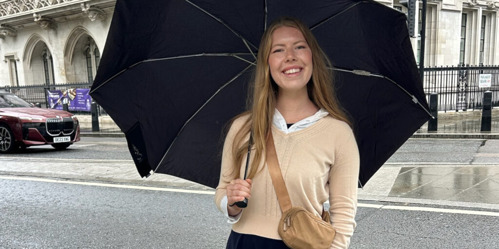 A top student and Union sophomore stands on a wet city street, smiling under a large black umbrella. She wears a beige sweater, crossbody bag, with buildings and a red car in the background.