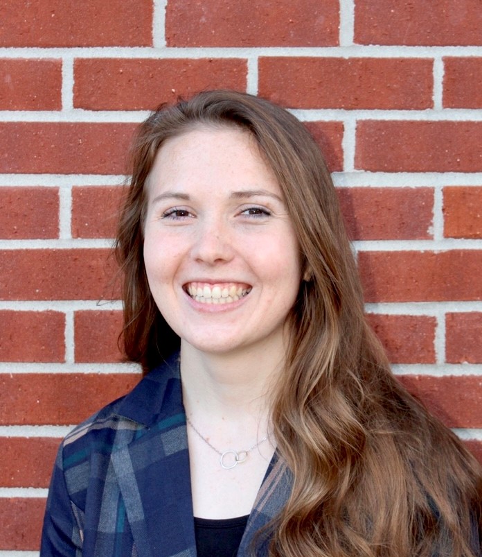 Taylor Overcast, a smiling woman with long brown hair stands in front of a red brick wall.